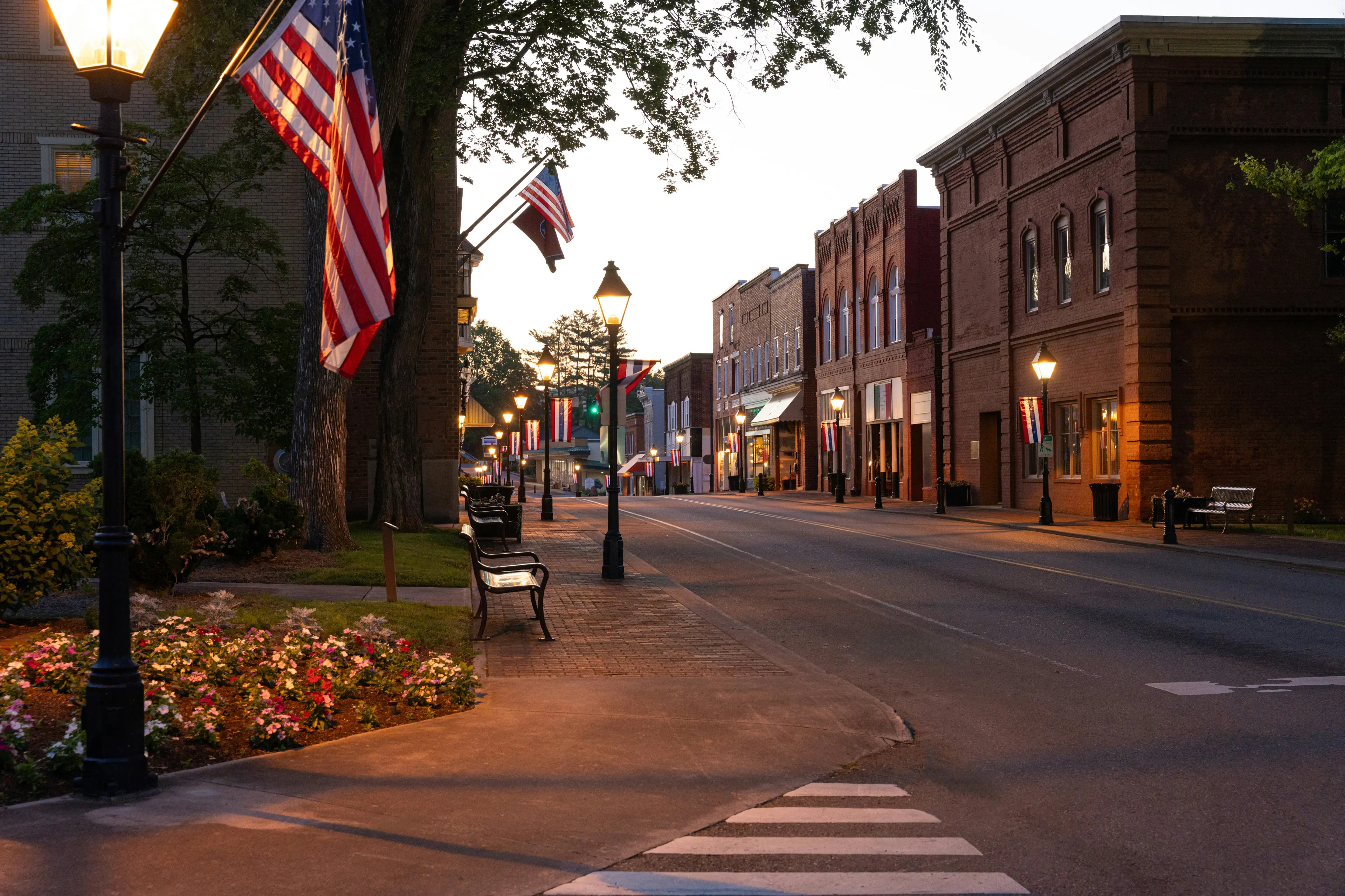 A small-town Main Street at dusk, flags on its lamp posts and lights on in its brick shopfronts